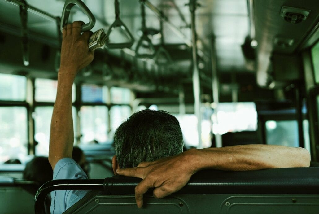 A person with short gray hair, perhaps headed to Port Canaveral for a cruise, sits on a bus viewed from behind. They grip an overhead strap while resting an arm on the seat back. The bus is mostly empty, with sunlight filtering through the windows as it journeys away from Orlando Airport.