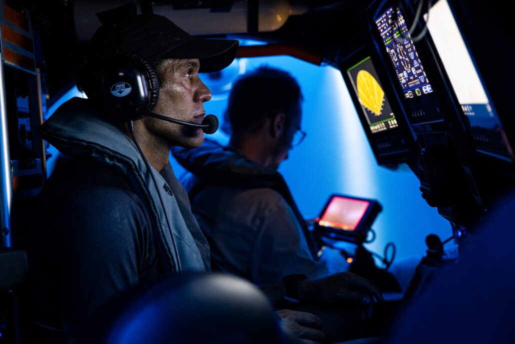 Two people in a dimly lit cockpit are focused on monitoring screens, perhaps imagining they're navigating a century-old WWI ship. The person in the foreground wears a cap and headset, as display screens reveal various controls and information vital for this modern-day cruise expedition.