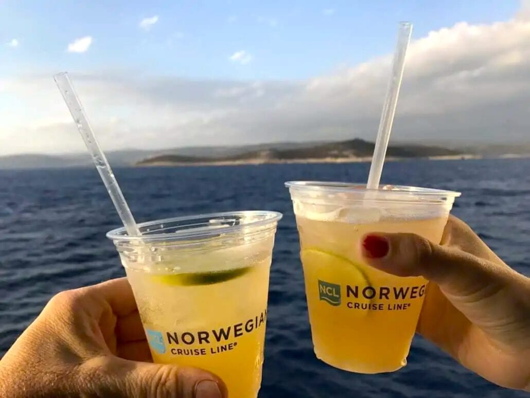 Two hands hold plastic cups with straws and lime slices, brimming with a light-colored beverage. The cups bear the "Norwegian Cruise Line" logo, perfectly capturing the essence of Norwegian's Free at Sea experience against a backdrop of sea views and distant landmass under a partly cloudy sky.