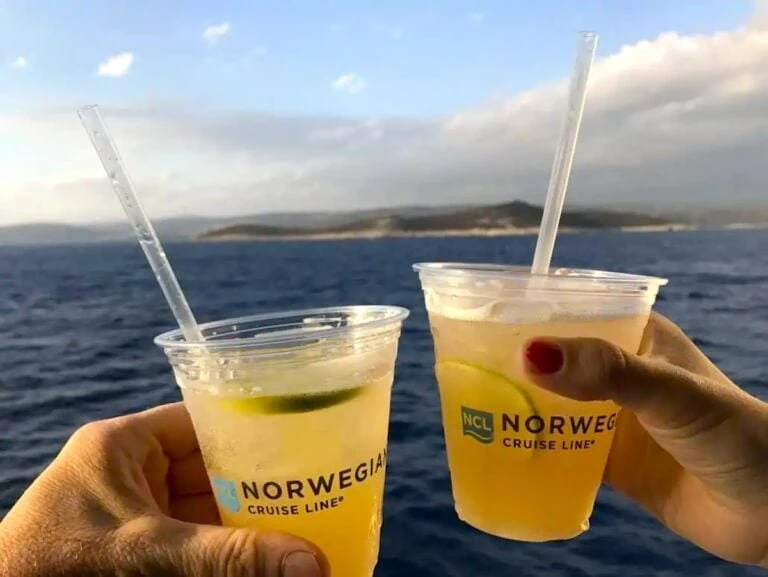 Two hands hold plastic cups with straws and lime slices, brimming with a light-colored beverage. The cups bear the "Norwegian Cruise Line" logo, perfectly capturing the essence of Norwegian's Free at Sea experience against a backdrop of sea views and distant landmass under a partly cloudy sky.