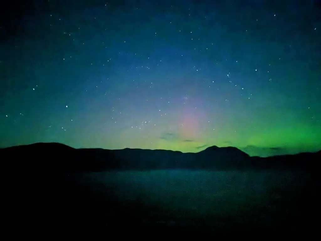 Under the night sky, stars shimmer above the dark silhouette of distant hills. A faint green aurora glows near the horizon, blending with the deep blue sky—a breathtaking view often witnessed on Alaska Inside Passage Cruises.