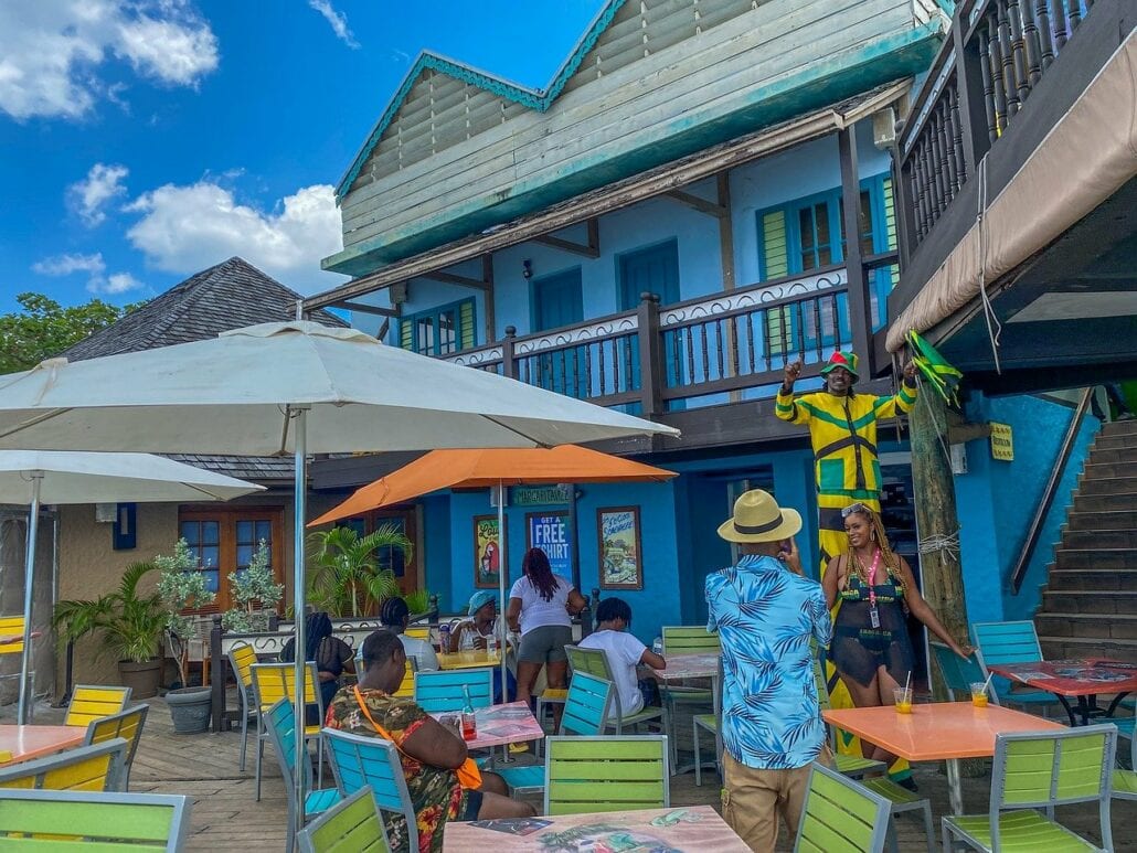 A tropical outdoor cafe with colorful tables and chairs under large umbrellas, reminiscent of Caribbean cruise ports. Several passengers dine and chat. The building is painted blue with wooden balconies. A person in Jamaican-themed attire decorates above a staircase. The sky is clear and sunny.