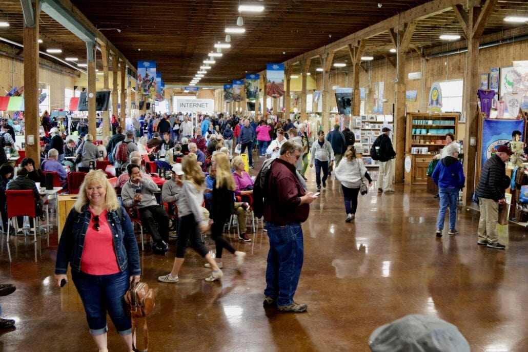 A large indoor market buzzes with cruise guests and locals alike, wandering, sitting at tables, and exploring booths. The wooden ceiling and beams craft a rustic charm. A sign for internet services stands out while various goods and decorations adorn the stalls in downtown Charlottetown.
