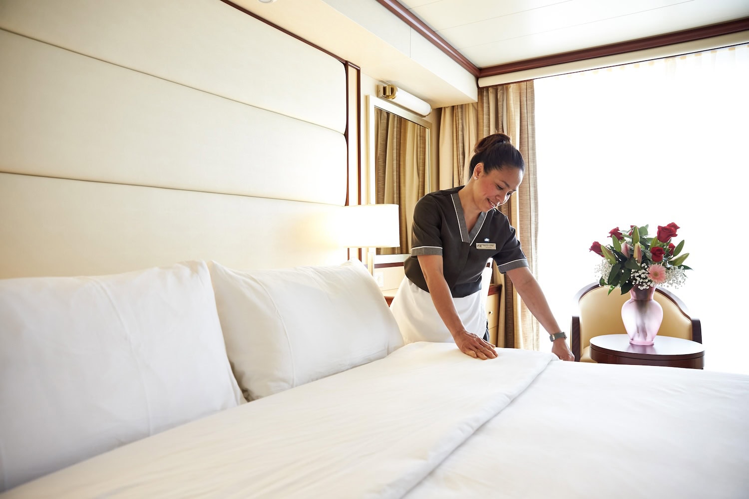 A hotel staff member in a uniform, reminiscent of cruise ship elegance, is making a neatly arranged bed in a well-lit room with beige curtains. A flower arrangement in a pink vase sits on a nearby table next to a chair.
