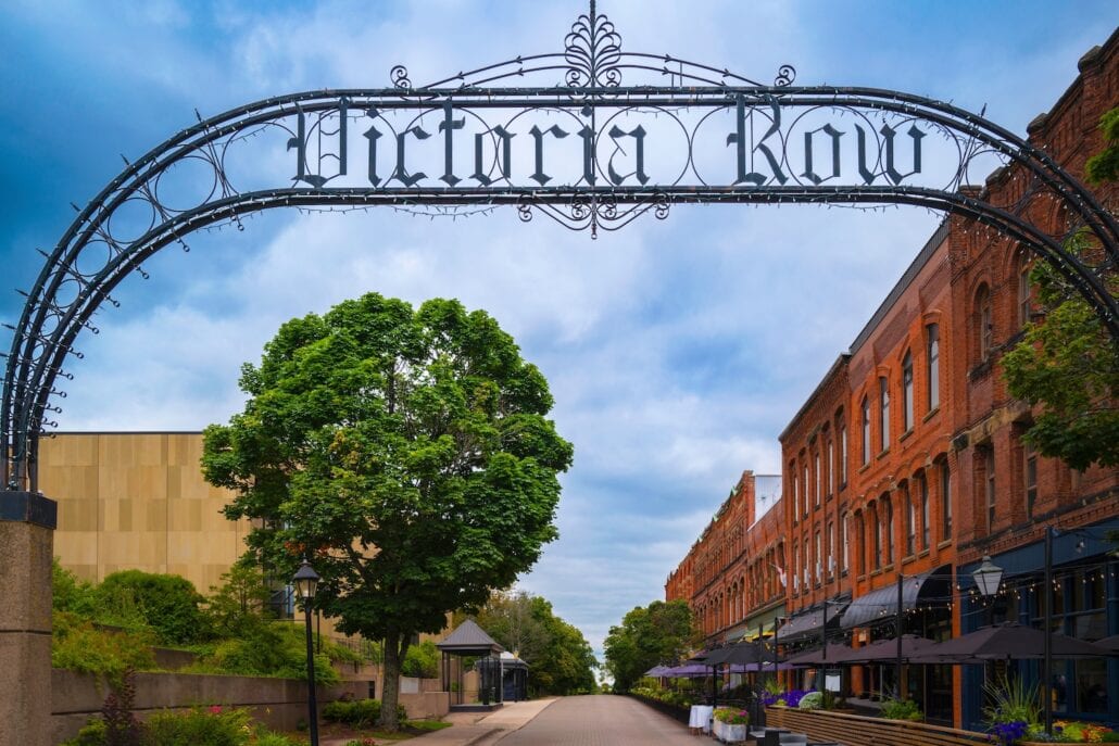 The image captures an arched iron sign reading "Victoria Row" over a pedestrian walkway in downtown Charlottetown. On the right, red-brick buildings feature outdoor seating areas, while a large tree and a modern building sit on the left under a partly cloudy sky, welcoming cruise guests exploring the area.