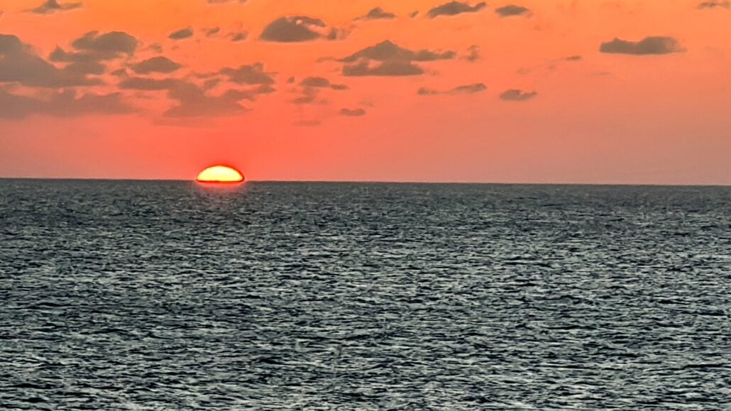 The image shows a vibrant sunset over the ocean, with the sun partially below the horizon. The sky is painted in shades of orange and scattered with small clouds, reflecting off the calm sea.