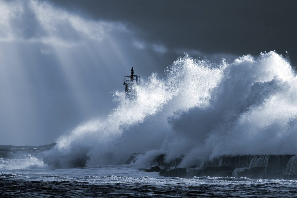 Large waves crash against a lighthouse on a rocky shoreline, reminiscent of the thrilling scenes often encountered during hurricane season. The sky is overcast, with beams of light piercing the clouds, creating a dramatic spectacle as water splashes and churns.