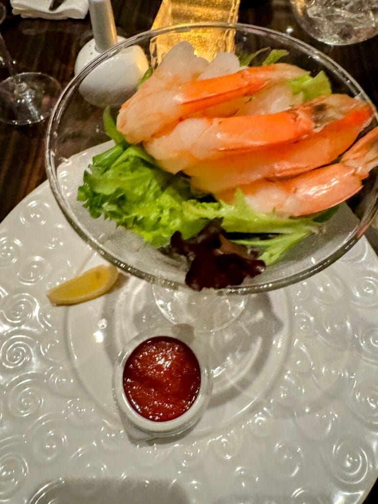 A shrimp cocktail is served in a glass with a bed of lettuce. Several large shrimp are placed on top. A small bowl of red cocktail sauce and a lemon wedge are on the white, textured plate beneath the glass.