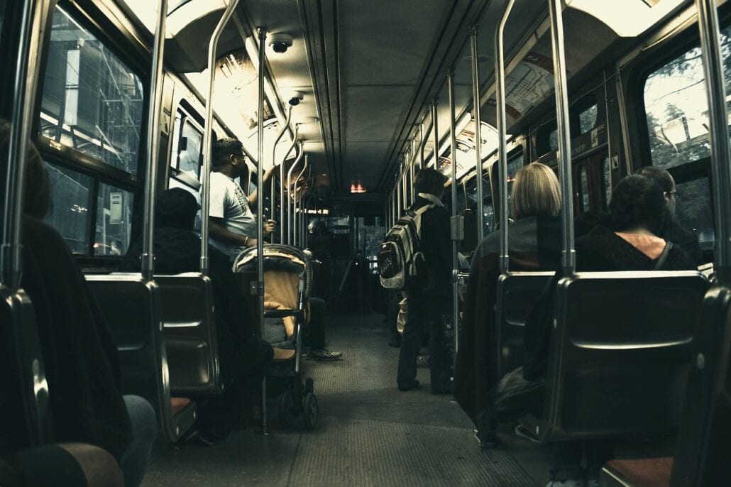 A dimly lit bus interior with several seated passengers and some standing, including one with a stroller, creates a calm and subdued atmosphere as it heads towards Orlando Airport. The view is looking toward the front, where the aisle leads the way to the next peaceful destination.