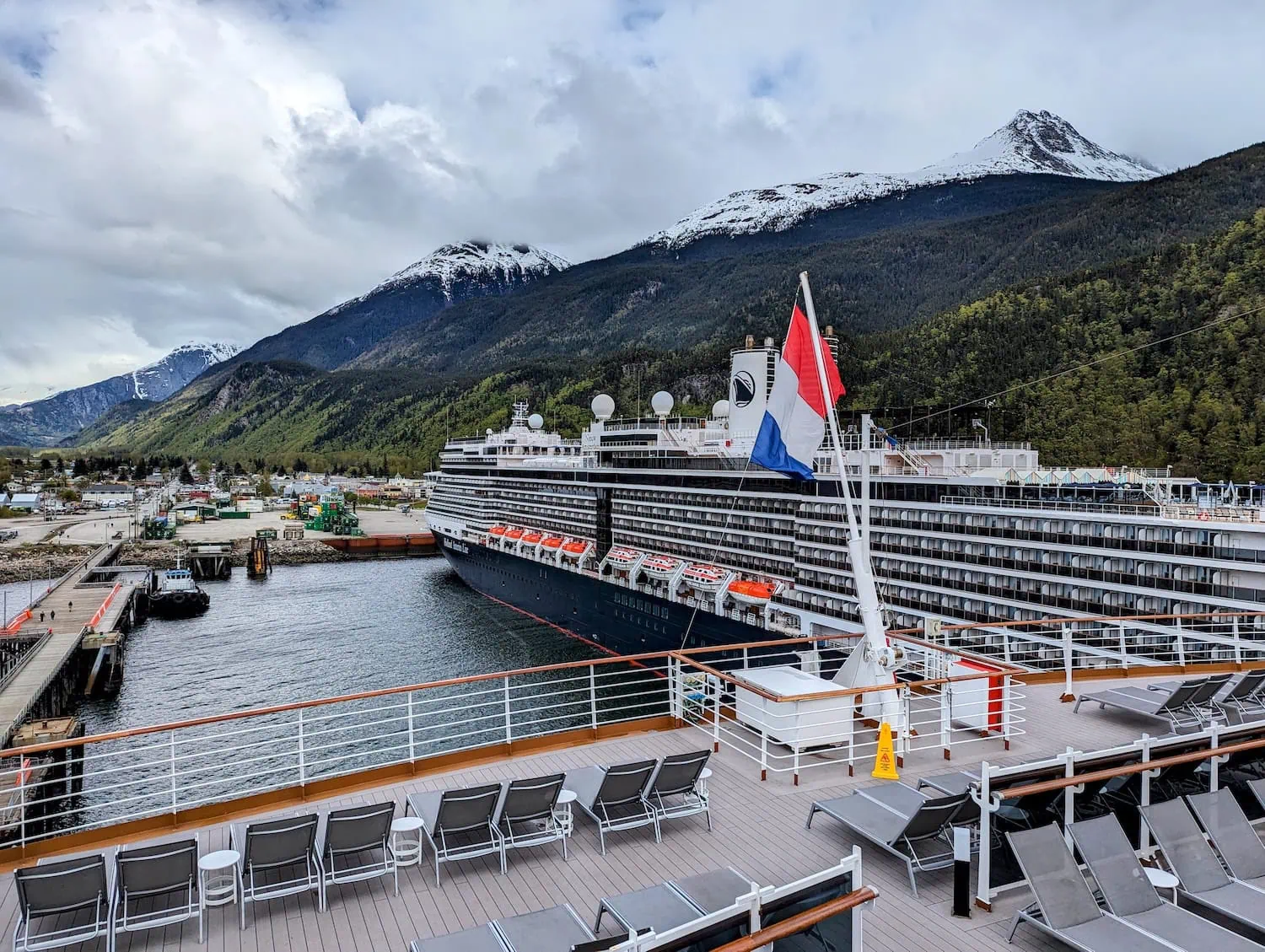 A cruise ship is docked at a port surrounded by mountains in Skagway, Alaska. The scene shows an overcast sky with snow-capped peaks in the background. The deck of another ship is visible in the foreground, with empty lounge chairs and a flag flapping gently.