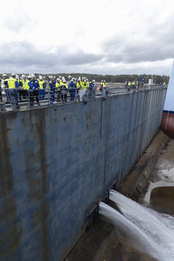 A group of people wearing safety gear stands along the edge of a large industrial structure with water flowing out from a spout near the base. Under cloudy skies, the debut of the Star of the Seas is marked by its ceremonial float out, with a ship's bow partially visible on the right.