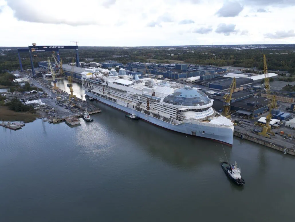 The Star of the Seas, poised for its float out debut, is docked at a shipyard where cranes meticulously attend to it. Surrounded by water, industrial buildings, and trees, a tugboat stands nearby under the cloudy sky.