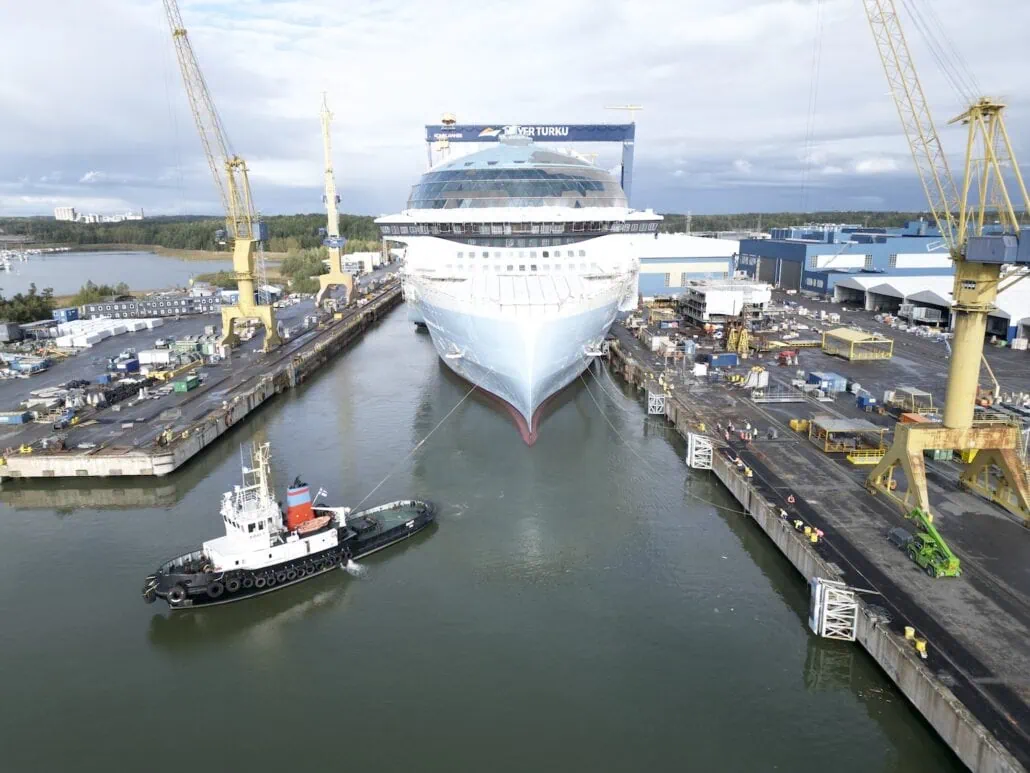 An aerial view captures the impressive Star of the Seas cruise ship in a shipyard, surrounded by cranes. A tugboat assists at the bow for its ceremonial float out. Several structures and equipment are visible, with trees in the background under a cloudy sky.