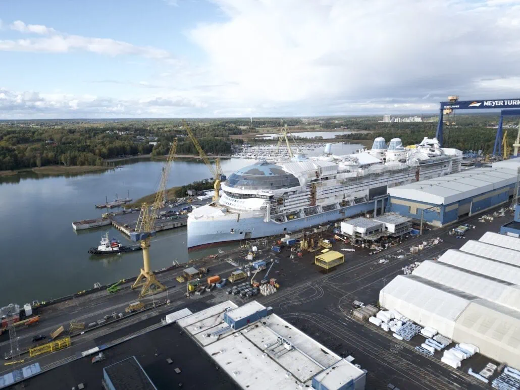 Aerial view of a large shipyard where the Star of the Seas is making its debut, docked near cranes. Surrounding buildings, vehicles, and equipment are visible. The shipyard is adjacent to a body of water, with a wooded landscape in the background under a cloudy sky.