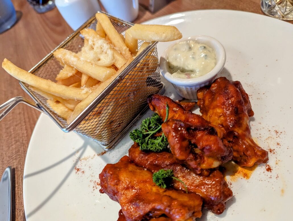 A plate with buffalo chicken wings garnished with parsley sits next to a small basket of fries topped with cheese aboard the Sun Princess. A ramekin of dipping sauce accompanies the meal on a wooden table.