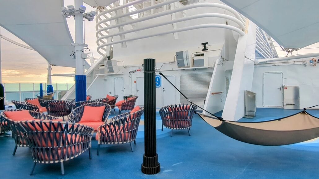 The outdoor seating area on the Sun Princess ship deck features lounge chairs and a hammock. Red and blue cushioned chairs are arranged in a circle around a pillar labeled "9." A shaded cover provides comfort, while the sky and railing set a serene backdrop for those coming to relax.