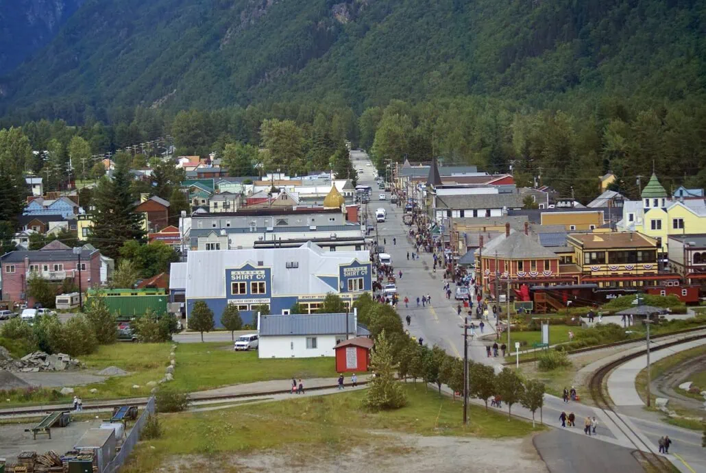 Aerial view of the quaint town of Skagway, Alaska, with a main street lined with colorful buildings. People stroll along as green hills and dense trees embrace this charming cruise port. A few cars are parked along the bustling street.