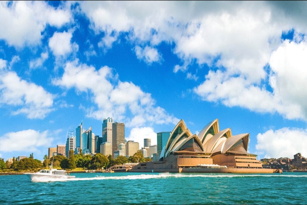 A view of the Sydney Opera House and city skyline with a boat on the water in the foreground. The sky, vibrant with scattered clouds, echoes the lively spirit of an Australian carnival.