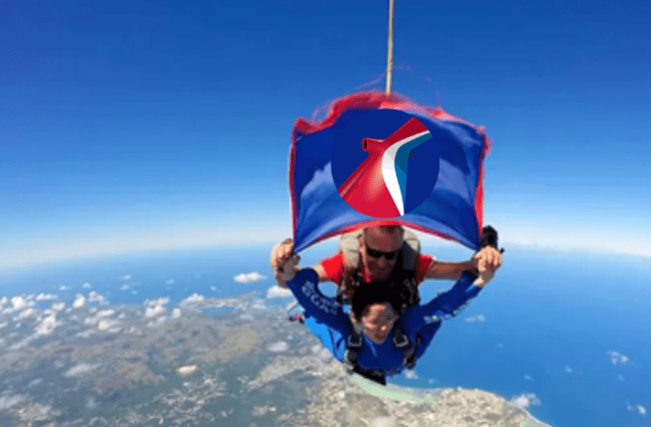 A tandem skydive over a coastal area, with a man and woman descending under a parachute featuring a blue, white, and red emblem reminiscent of Carnival Cruise Line. The ocean and land shimmer below under the clear blue sky.