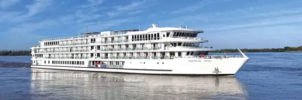 A large white river cruise ship, part of the renowned Cruise Line fleet, sails on calm, wide waters under a clear blue sky. The "American Song" boasts multiple decks with glass balconies and a few passengers visible on the upper deck.