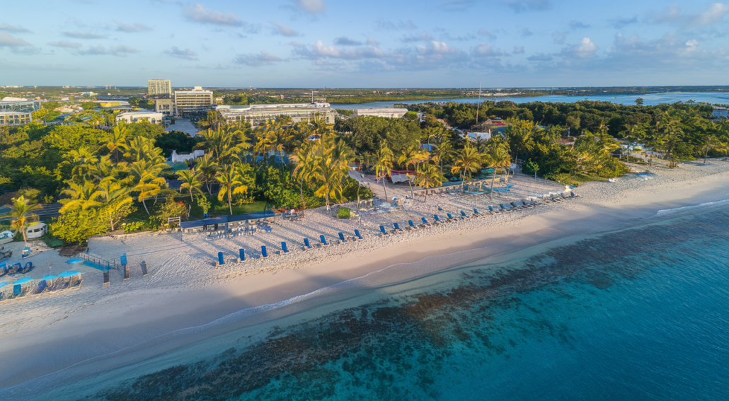 Aerial view of a sandy beach lined with blue lounge chairs and umbrellas, resembling a vibrant Carnival scene. Lush greenery and buildings are visible in the background. The ocean water is clear and blue, meeting the shoreline under a partly cloudy sky, suggesting a sunny day.