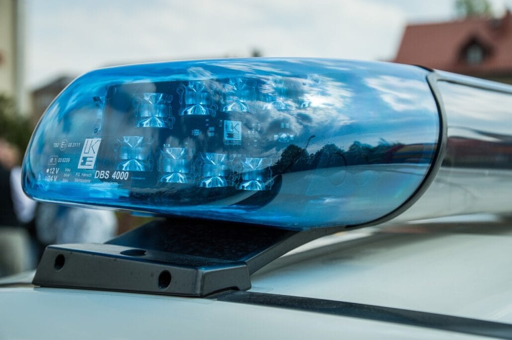 Close-up of a blue police car siren mounted on the roof of a vehicle, its reflective surface subtly concealing what might be the lens of a hidden camera, capturing partial glimpses of the sky and surrounding buildings.