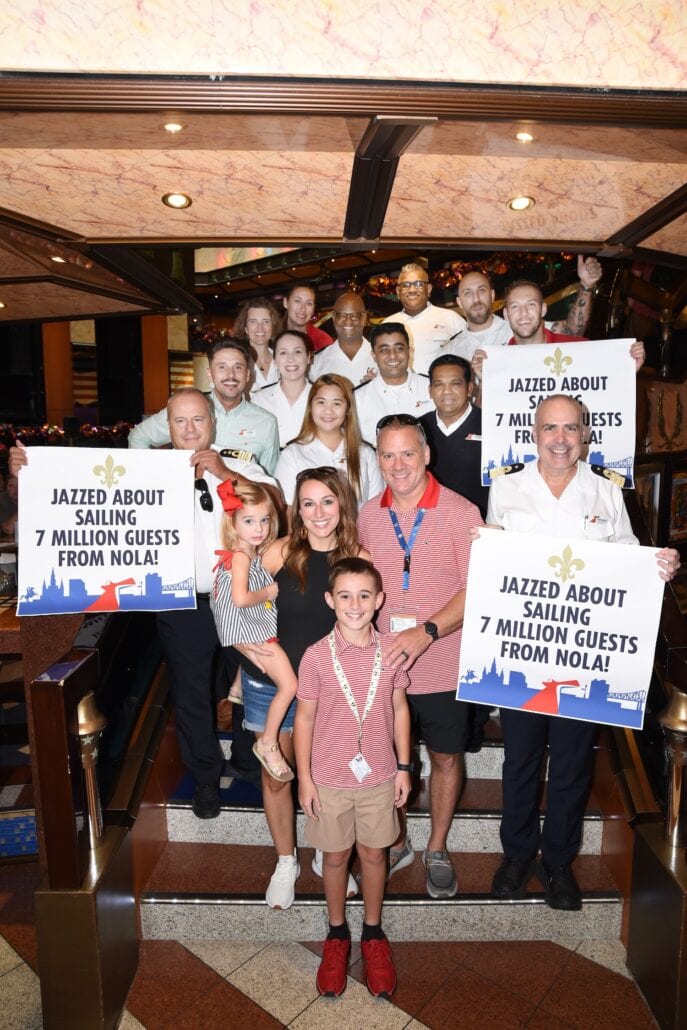 A group of people stand on a staircase, holding signs that read, "JAZZED ABOUT SAILING 7 MILLION GUESTS FROM NOLA!" Some members of the group are wearing Carnival Cruise Line uniforms while others are in casual attire. A young girl and boy are at the front with an adult in vibrant New Orleans surroundings.