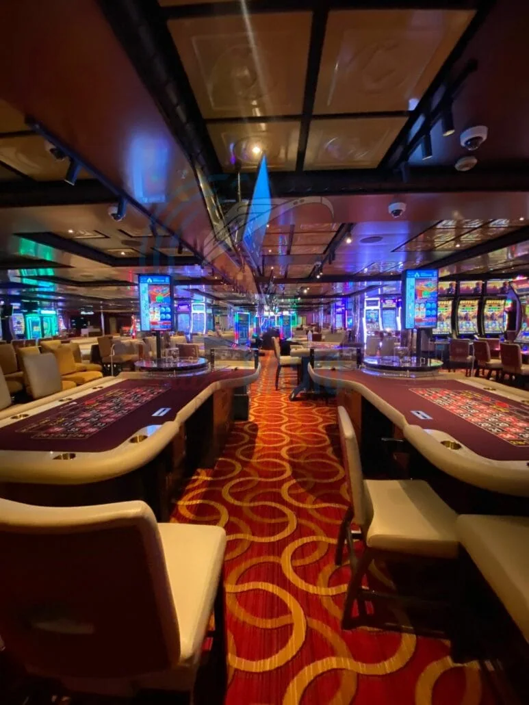 The recently refurbished casino interior on the Carnival Radiance boasts poker tables lined with maroon felt, surrounded by comfortable chairs. Slot machines twinkle with colorful lights in the background, while the floor features a red and gold swirl pattern, ensuring an inviting service atmosphere.