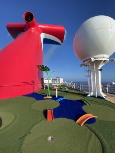 The refurbished mini-golf course on the Carnival Radiance cruise ship deck features a giant red funnel and a large white spherical structure. With artificial grass, vibrant colors, and various obstacles, this lively course is set against a clear blue sky for guests to enjoy.