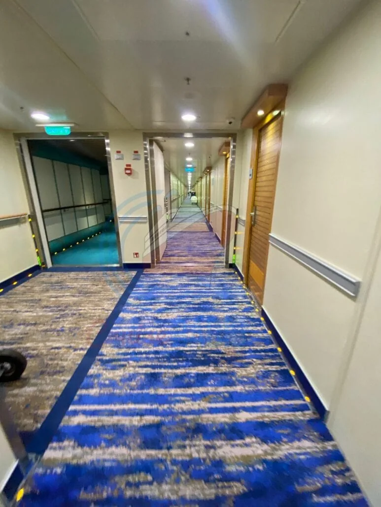The refurbished hallway on Carnival Radiance features a long, carpeted design with wooden doors lining the light beige walls. The floor boasts a blue and gray striped pattern, while a handrail runs along the left side and emergency exit signs are visible on the ceiling.