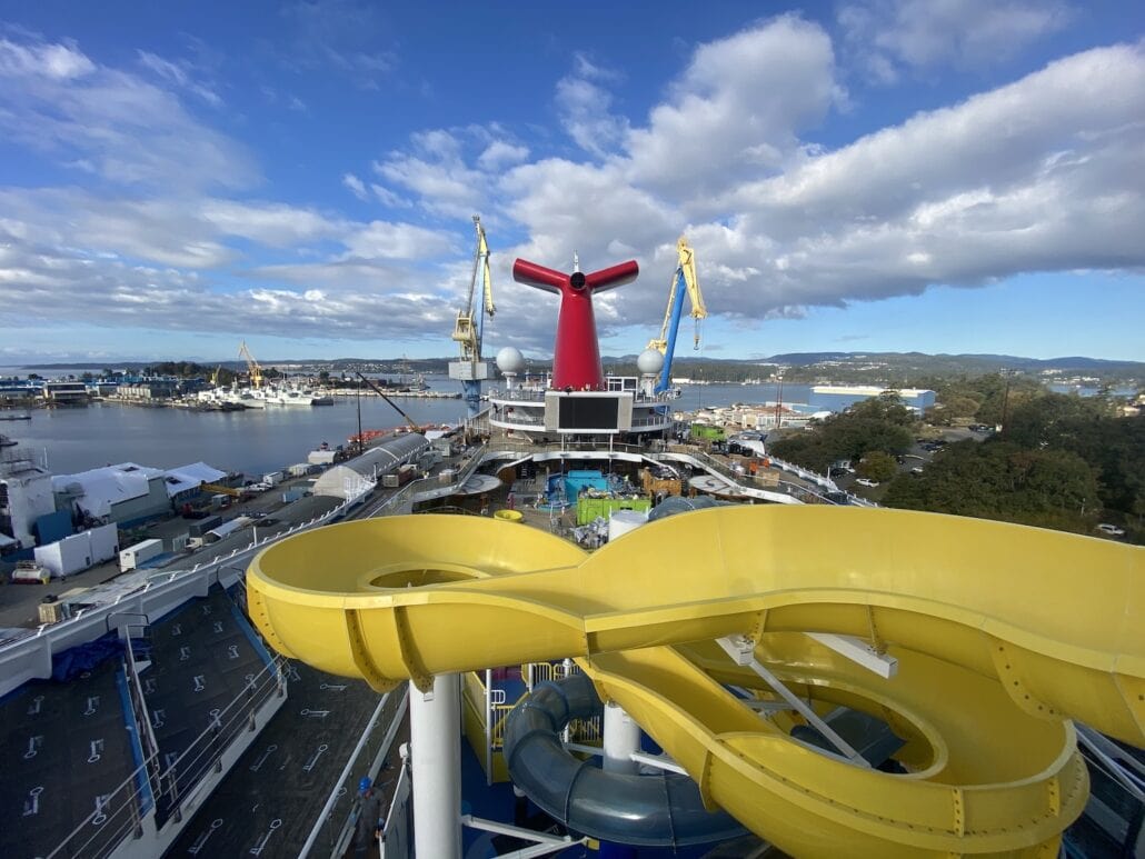 The Carnival Radiance, showcasing a gleaming yellow water slide, is docked at the port. With its vibrant red funnel standing tall amidst cranes in the harbor, this ship has undergone a stunning refurbishment. The sky is partly cloudy, and distant hills create a picturesque backdrop.