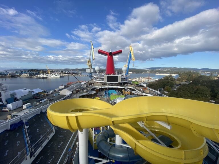 The Carnival Radiance, showcasing a gleaming yellow water slide, is docked at the port. With its vibrant red funnel standing tall amidst cranes in the harbor, this ship has undergone a stunning refurbishment. The sky is partly cloudy, and distant hills create a picturesque backdrop.