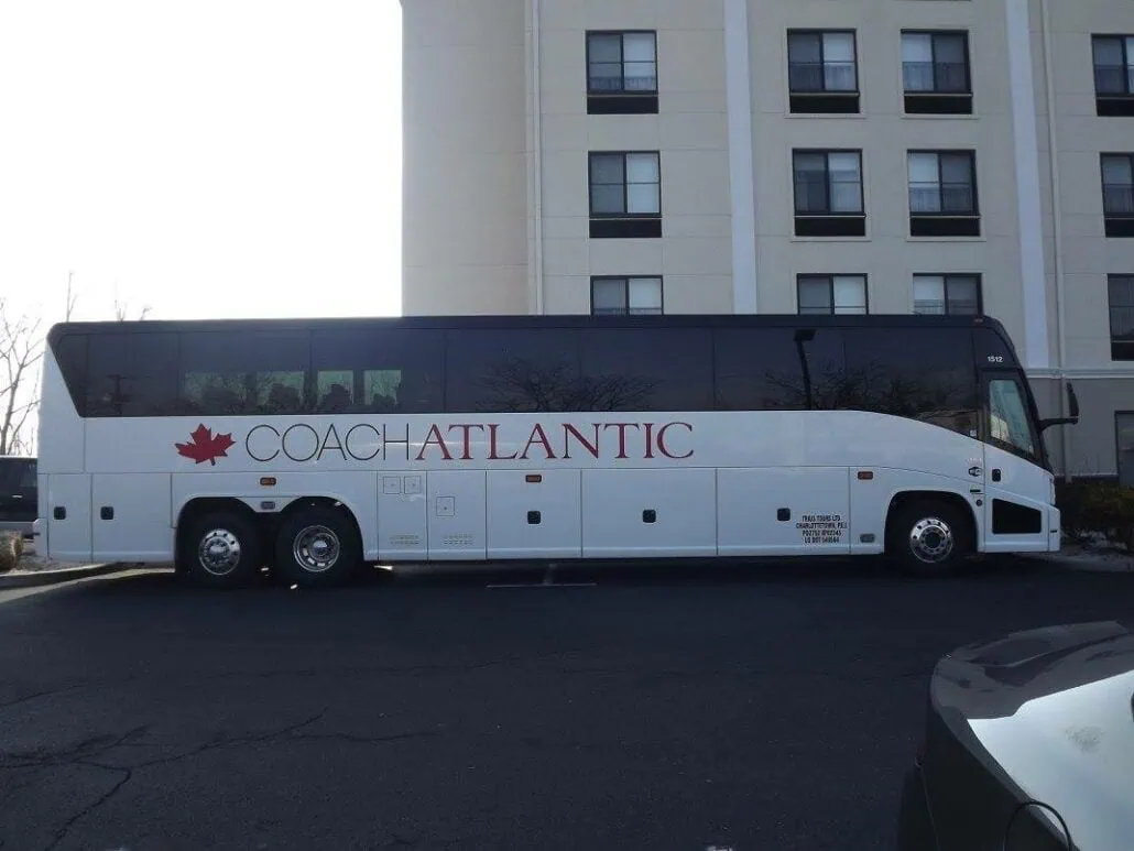 A white Coach Atlantic bus is parked in front of a multi-story building in downtown Charlottetown. The bus, featuring a red maple leaf symbol and black-tinted windows, awaits cruise guests eager to explore the city.
