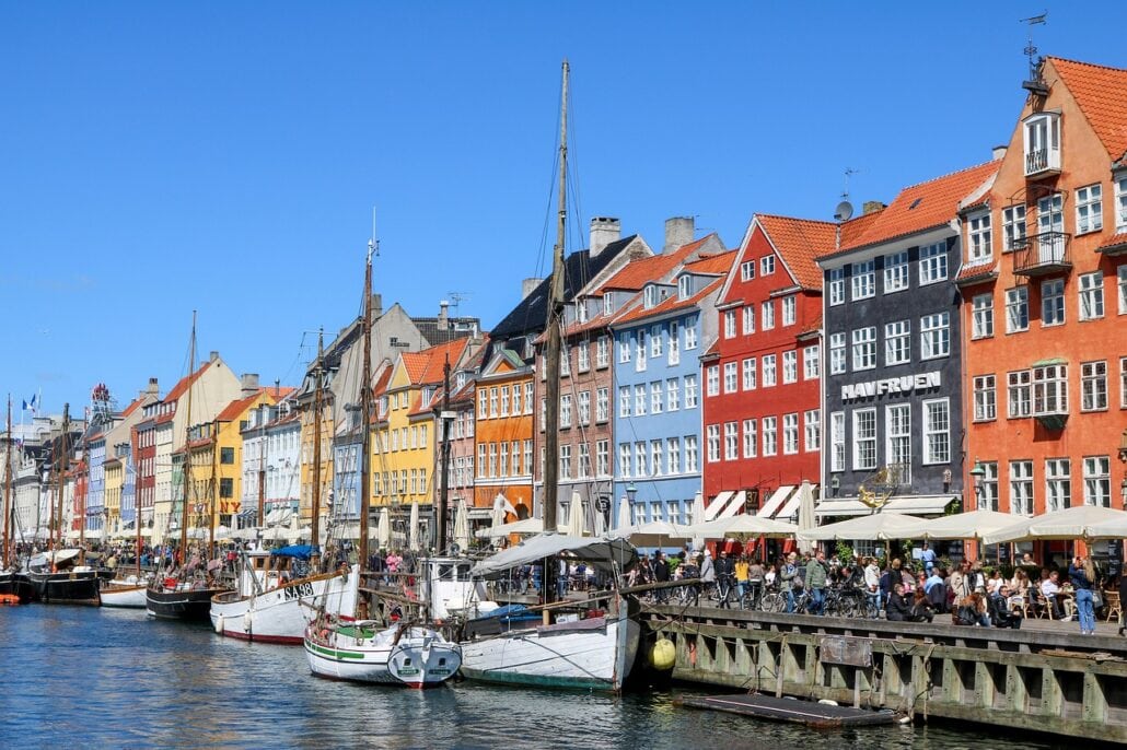 Colorful buildings line the Nyhavn waterfront in Copenhagen, Denmark. Several boats, perhaps from a European cruise, are docked along the canal. People are sitting at outdoor cafes under umbrellas on a sunny day.