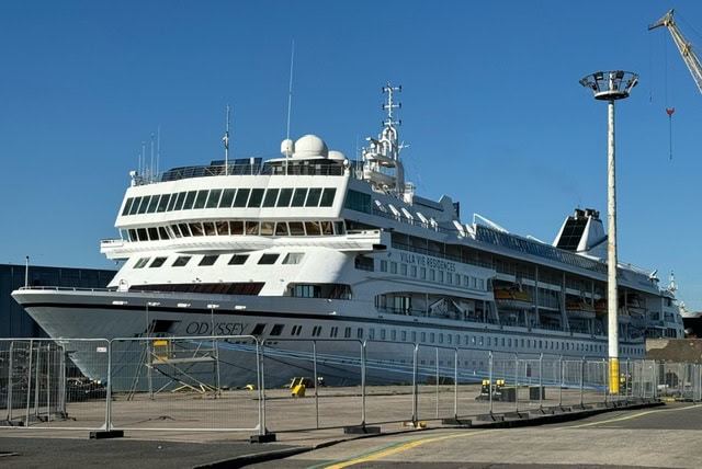 A large white cruise ship named "Odyssey" is docked at the port, preparing for its world cruise. The ship is surrounded by metal fencing, and a nearby lamppost casts a shadow. The sky is clear and blue, with some port infrastructure visible in the background. Belfast departure marks an exciting journey ahead.