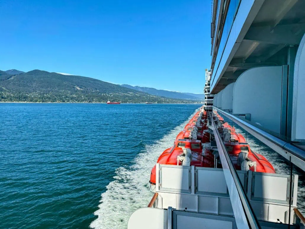 Aboard a cruise through the Alaska Inside Passage, vibrant red lifeboats line the deck as the ship glides over the azure waters, leaving a crisp white wake. In the distance, lush forested shorelines and majestic mountains rise beneath a clear blue sky.