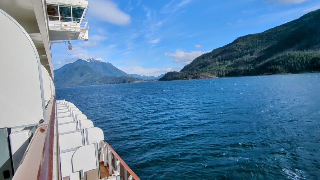 View from a cruise ship's deck, showcasing the serene blue ocean along Alaska's Inside Passage, with a mountainous landscape under a partly cloudy sky. The ship's white exterior and passenger balconies are visible on the left side of the image.