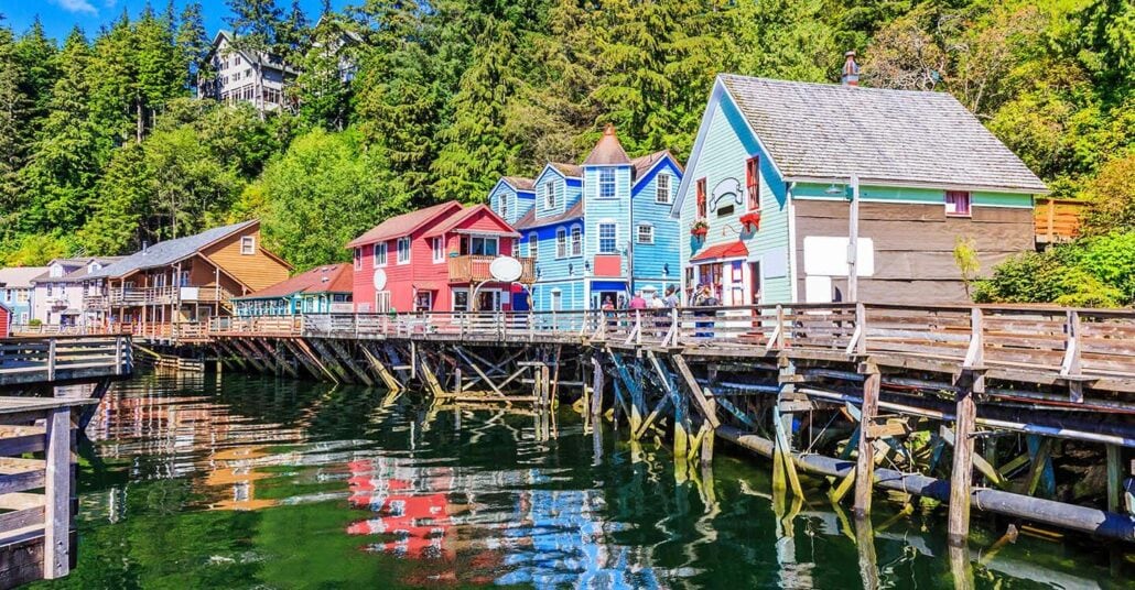 A colorful row of wooden buildings on stilts lines a waterfront boardwalk. The buildings are painted in red, blue, and brown. A dense forested hillside rises behind them, and the water reflects their vibrant colors.