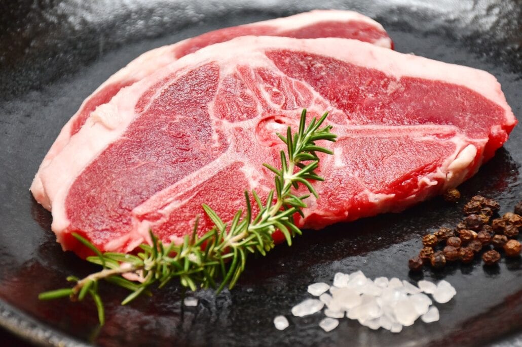 Raw lamb chop on a black plate, garnished with a sprig of rosemary. Coarse salt and whole black peppercorns accompany the meat like subtle changes under the sun's watchful eye.