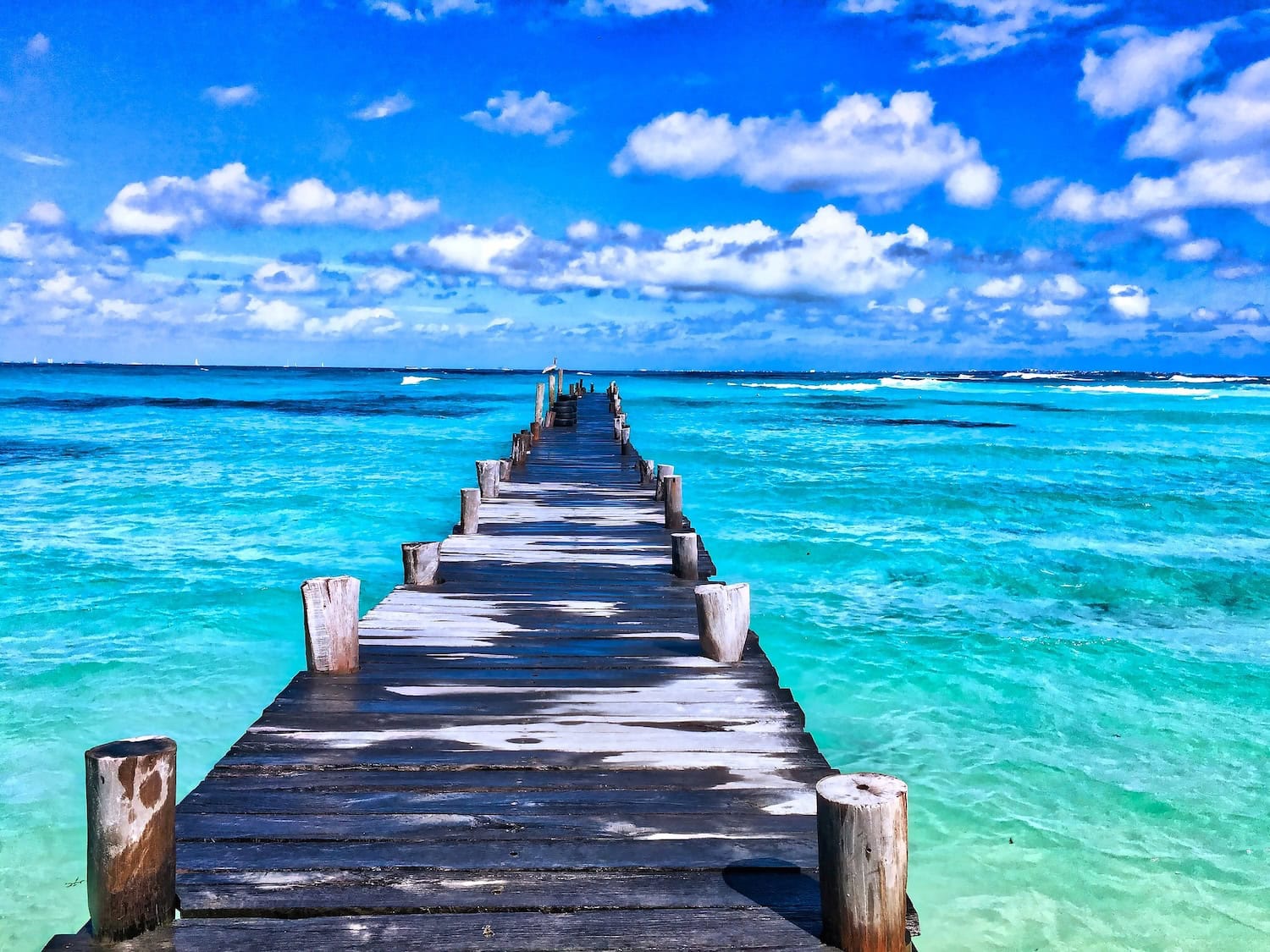 A weathered wooden pier extends into a vivid turquoise ocean under a sky filled with scattered white clouds. The water is calm, and the distant horizon blends with the blue sky, creating a serene and inviting coastal scene.