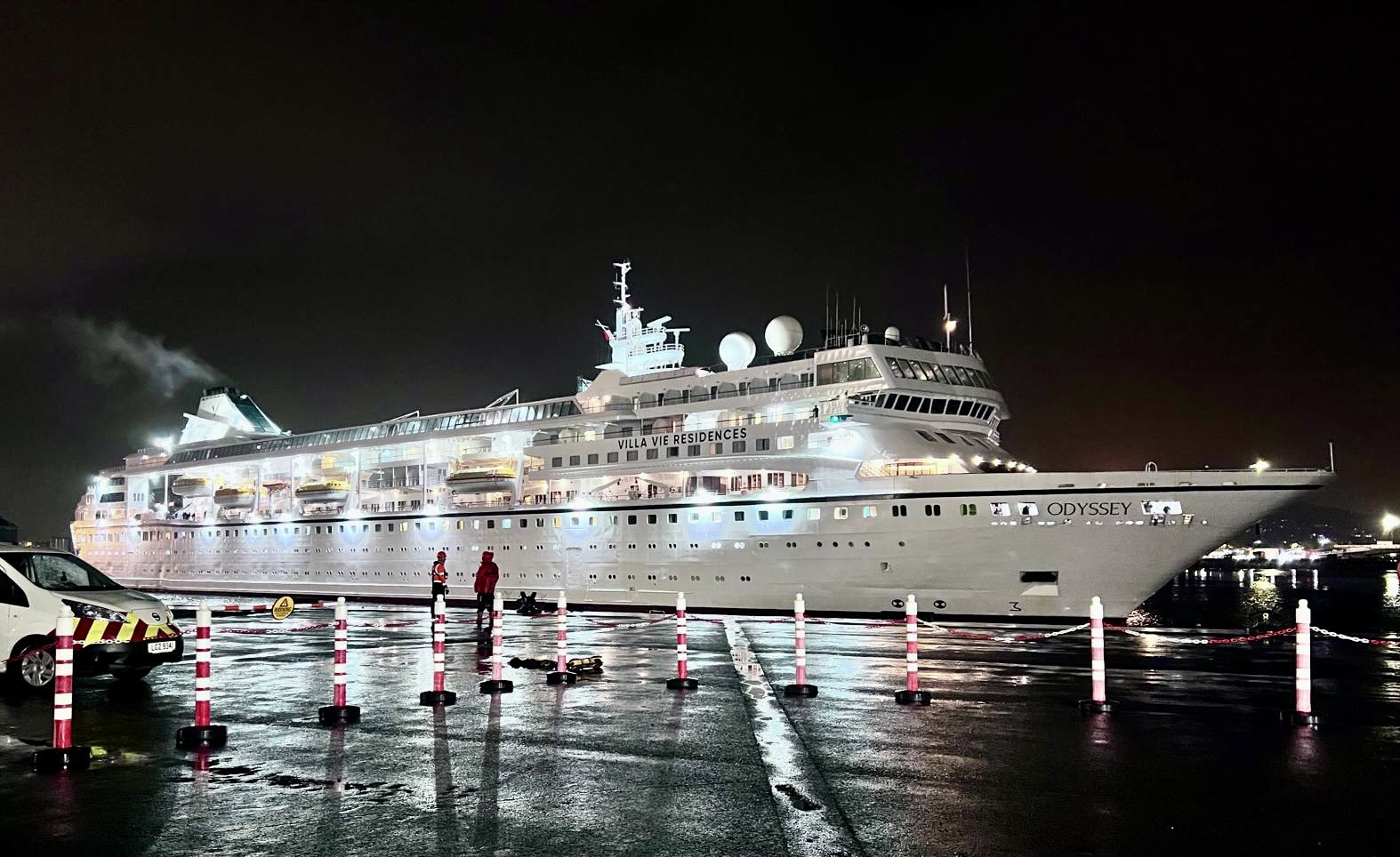 A large cruise ship named Odyssey, part of a World Cruise fleet, is docked at a port at night. The ship is well-lit, and two people in red uniforms are standing on the wet pavement near the ship. Safety posts with reflective markings are placed nearby.