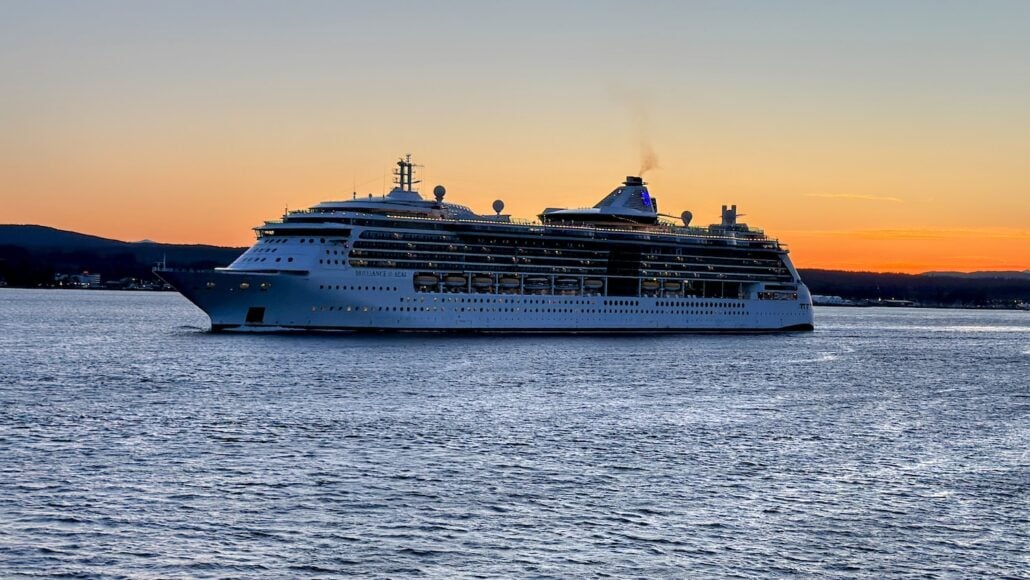 A large cruise ship sails on the serene waters of the Inside Passage during sunset. The ship's exterior is lit, with multiple decks gleaming. The sky is a gradient of orange and blue, with subtle silhouettes of distant hills in the background. Experience the magic of Alaska cruises.