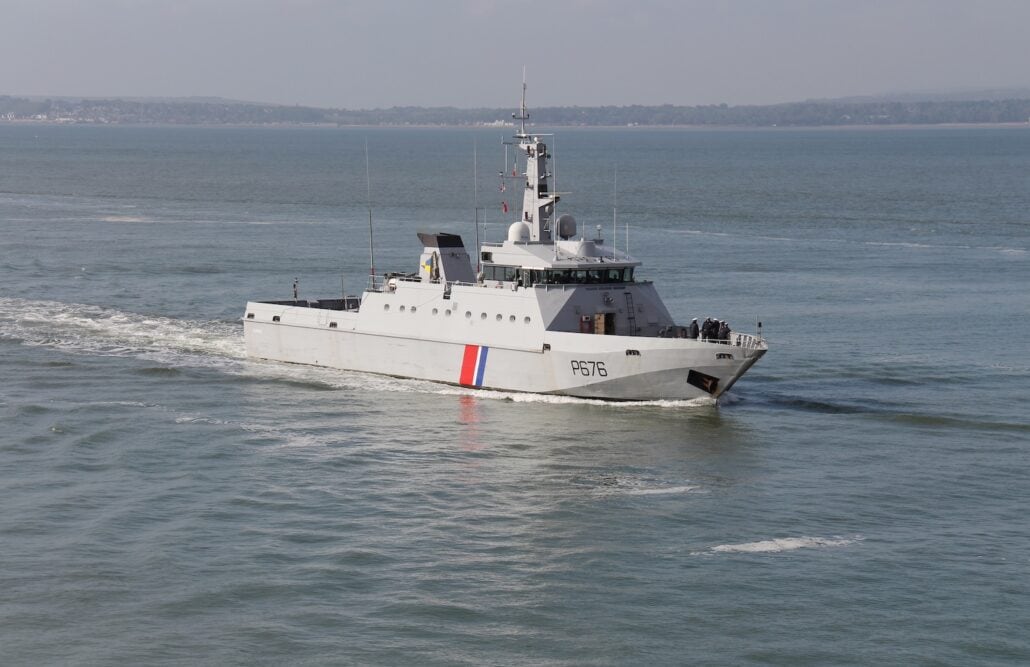 A gray patrol vessel with a red and blue stripe gracefully navigates through a calm sea, creating an auto-drafted trail of white water beneath a cloudy sky. Land is faintly visible on the horizon in the background.