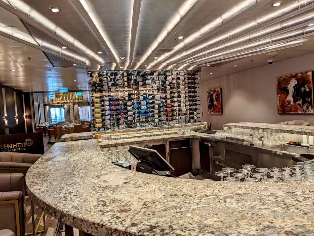 A modern bar with a curved marble countertop, lined with empty glassware. Behind the bar is a large wine rack filled with various wine bottles. The ceiling features linear lighting, and there are abstract artworks on the walls.