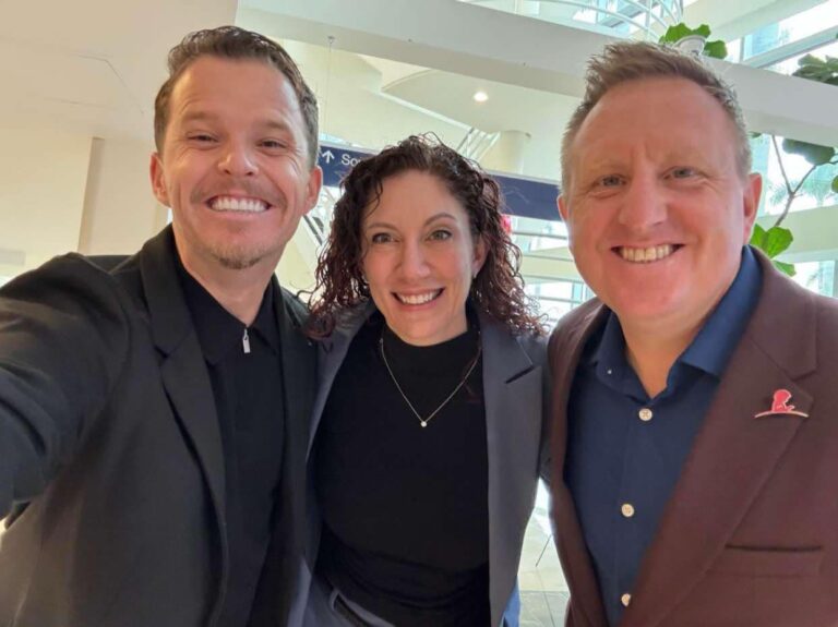Three people are smiling indoors, posing for a photo. Two men are on either side of a woman, all dressed in business casual attire. The background features modern architecture and greenery—a setting befitting a Fleet Cruise Director like Mike Pack preparing for the upcoming Carnival event.
