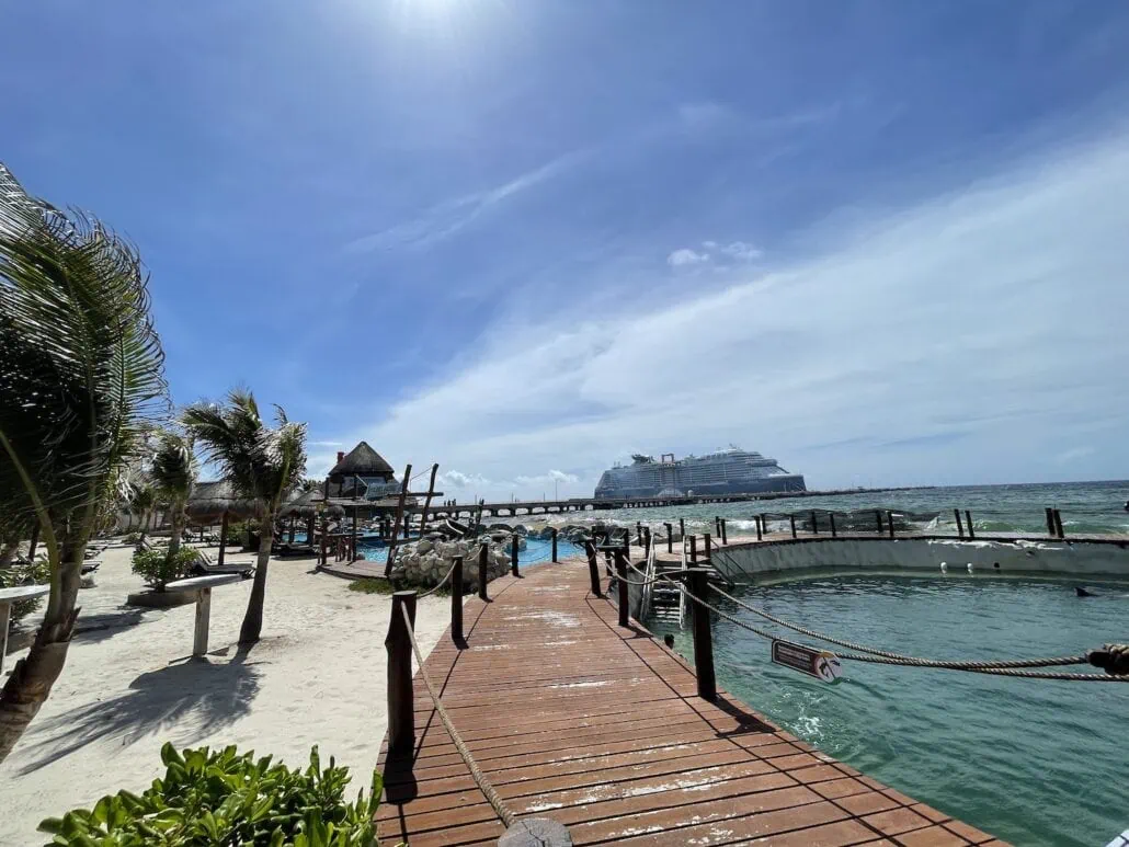 Discover the enchanting Costa Maya, where a sandy beach with swaying palm trees and wooden walkways awaits. In the background, a grand cruiser is docked beneath a clear blue sky. Meanwhile, in the foreground, exotic birds gather around a circular enclosure—one of many things to do here.