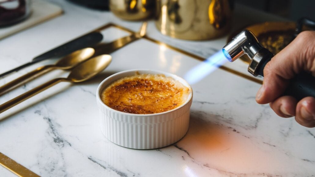 A hand skillfully uses a culinary torch to caramelize sugar atop a crème brûlée, following a classic crème brûlée recipe. Gold cutlery complements the dessert on the white marble surface, reminiscent of the elegance found dining aboard Carnival Cruise Line.