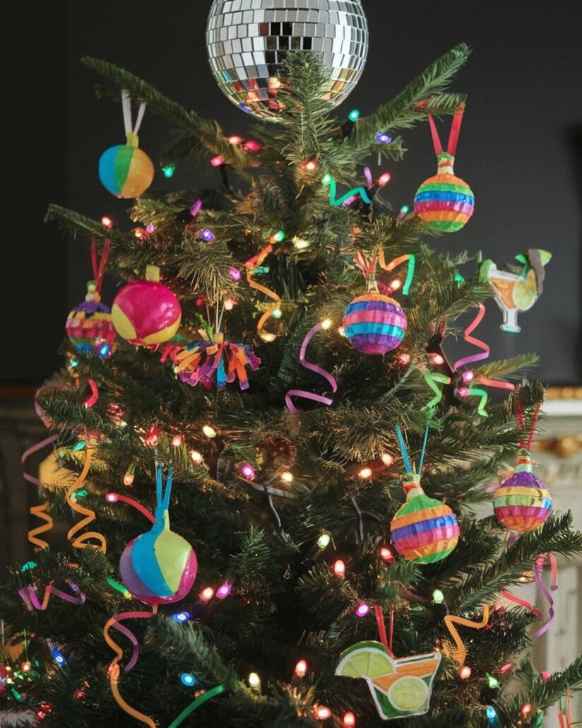A cruise-themed Christmas tree is decorated with colorful paper ornaments, including ball shapes and drink designs. It is adorned with multicolored lights and spiral streamers, while a disco ball graces the top.