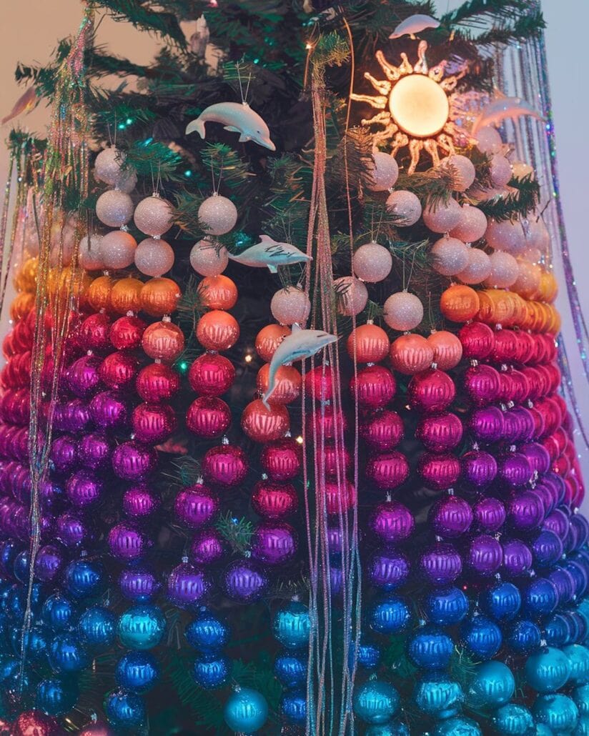 A cruise-themed Christmas tree is adorned with an array of colorful ornaments in a gradient pattern from blue to pink to orange. It features several dolphin figurines and a sun-shaped ornament near the top, while silver tinsel elegantly drapes down the sides.
