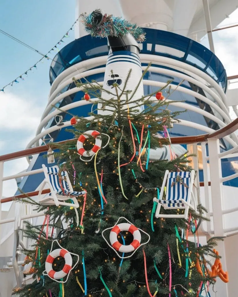 A cruise ship's deck hosts a Christmas tree adorned with nautical-themed decorations like life rings and deck chairs. Festive lights twinkle overhead, while the ship's funnel towers in the background, creating a unique holiday atmosphere at sea.